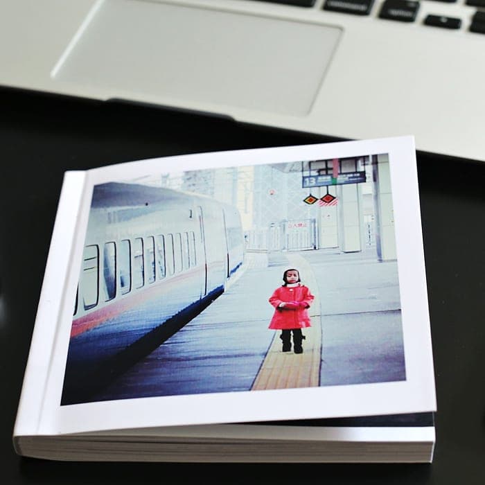 A 5×5 photobook resting on a desk, its cover a small child in a red coat waiting on a train platform.
